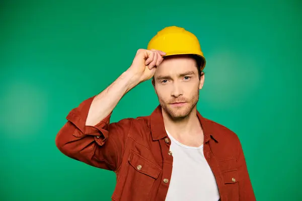 Un trabajador masculino guapo en uniforme y herramientas luciendo un sombrero amarillo sobre un fondo verde. - foto de stock