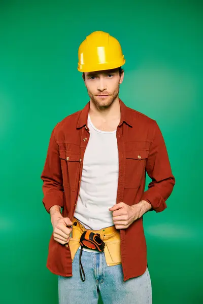 Un trabajador masculino guapo en uniforme y sombrero duro se para con las manos en los bolsillos sobre un fondo verde. - foto de stock