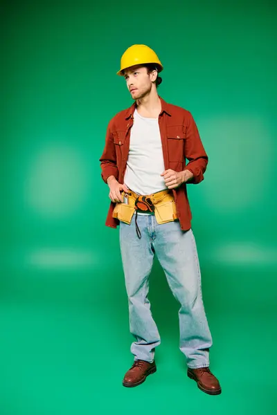 Un hombre con un sombrero duro sosteniendo un cinturón de herramientas, listo para trabajar en un fondo verde. - foto de stock