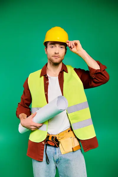 Trabajador guapo en sombrero duro y chaleco de seguridad posando con herramientas sobre fondo verde. - foto de stock
