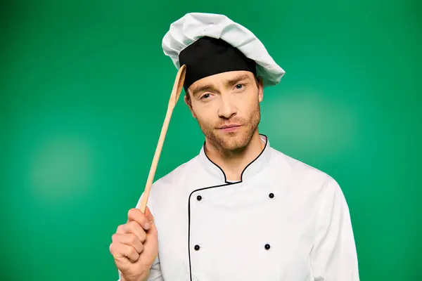 A handsome male chef in white uniform holding a wooden spoon against a green backdrop. — Stock Photo