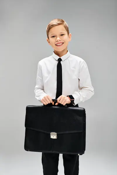 Чарівний хлопчик у білій сорочці і краватці тримає чорний портфель на сірому тлі. — стокове фото