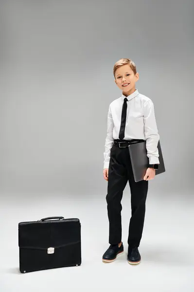 Un ragazzo elegante si trova con fiducia accanto a una valigetta su uno sfondo grigio. — Foto stock