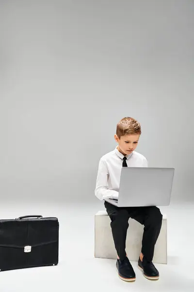Little boy in smart attire sits on stool absorbed in laptop work, against gray backdrop. — Stock Photo