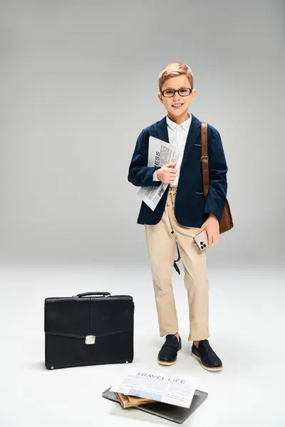 Preadolescent boy in elegant attire stands next to a briefcase against a gray backdrop. — Stock Photo