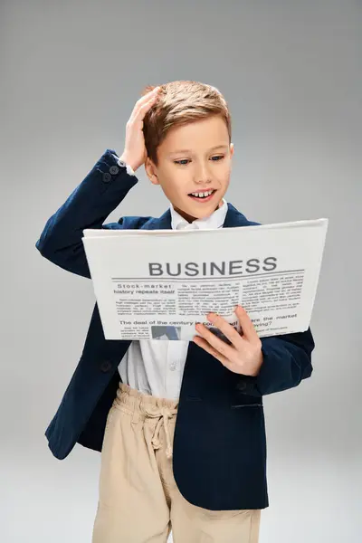 Elegant preadolescent boy in suit reading newspaper on gray backdrop. — Stock Photo