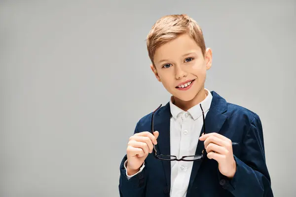 Niño en traje elegante sostiene un par de gafas contra un telón de fondo gris. - foto de stock