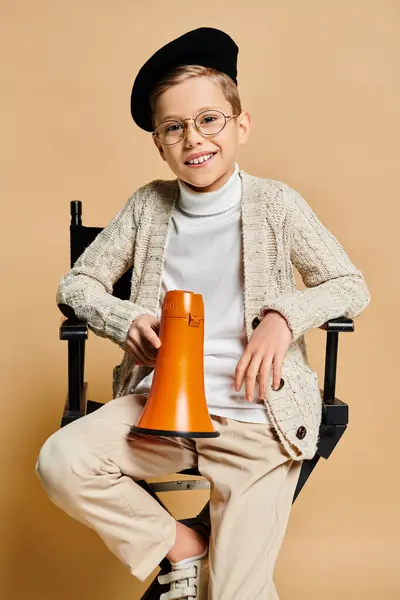 Jeune garçon habillé en réalisateur, tenant un mégaphone orange assis sur une chaise. — Photo de stock