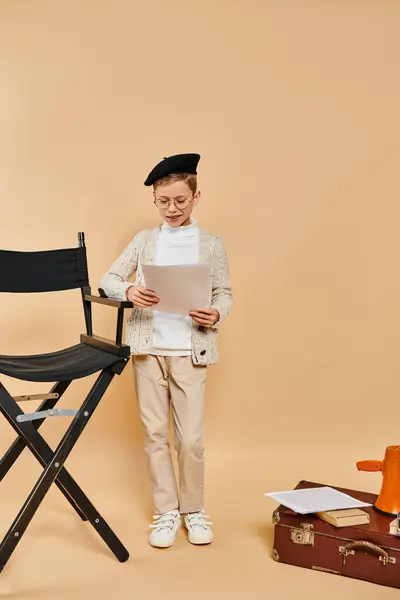 Preadolescent boy in film director costume holds paper next to chair. — Stock Photo
