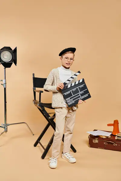 A preadolescent boy in film director attire holds a movie clapper in front of a camera. — Stock Photo