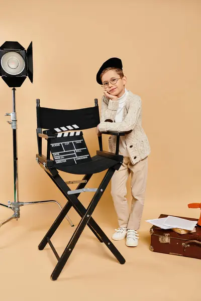 Un ragazzo preadolescente vestito da regista si trova con fiducia accanto a una sedia da regista. — Foto stock
