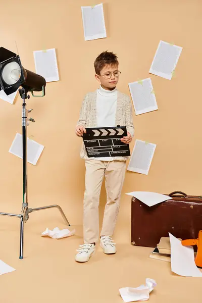 Preadolescent boy holds movie clapper in front of camera on beige backdrop. — Stock Photo