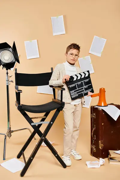 Ein vorpubertärer Junge in Filmregisseurskleidung hält einen Filmklöppel neben einem Stuhl. — Stockfoto