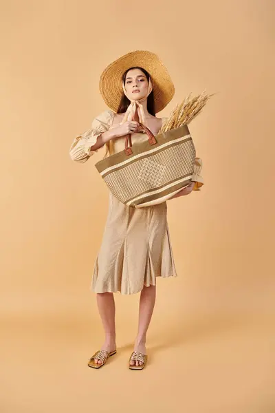 Une jeune femme aux longs cheveux bruns pose élégamment dans un studio, portant un chapeau et tenant un sac avec une humeur estivale. — Photo de stock