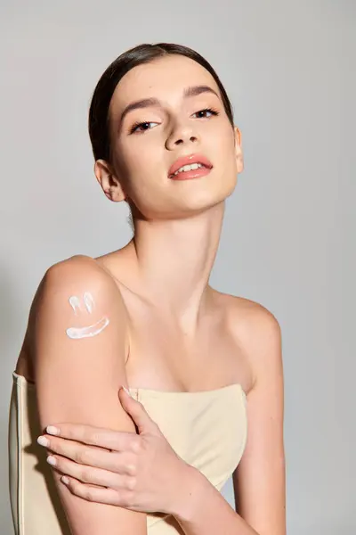 Une jeune femme aux cheveux bruns a l'air radieuse dans une robe blanche alors qu'elle pose avec une crème sur le bras dans un décor de studio. — Photo de stock