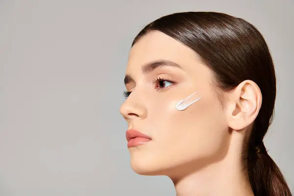A young woman with brunette hair applying cream on her face, exuding confidence and self-love in a serene studio setting. — Stock Photo