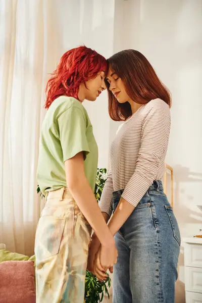 Serene young lesbian couple with closed eyes holding hands while standing at home, bliss and love - foto de stock