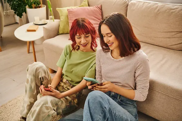 Heureux couple lesbien souriant et en utilisant des smartphones près du canapé, mode de vie moderne et les médias sociaux — Photo de stock