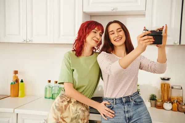 Young lesbian couple smiling and taking selfie on retro camera in kitchen, capturing blissful moment — Stock Photo