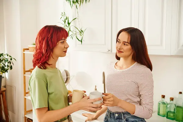 Feliz pareja lesbiana compartiendo una bebida caliente mientras sostiene tazas en su cocina llena de luz - foto de stock