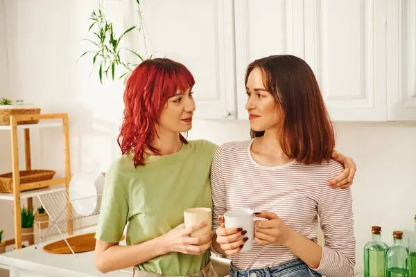 Happy lgbt couple sharing a warm beverage while holding cups in their kitchen filled with light — Stock Photo
