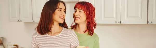 Happy lesbian couple sharing a warm hug while standing in kitchen filled with light, banner — Stock Photo