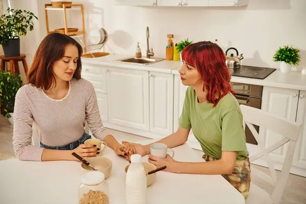 Acogedor momento de una joven pareja de lesbianas cogidas de la mano durante el desayuno en una acogedora cocina - foto de stock