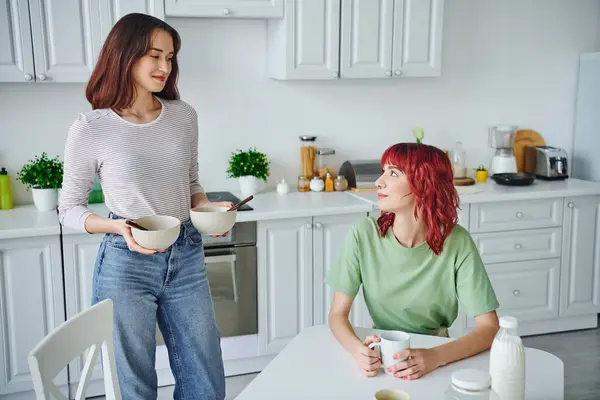 Heureuse jeune femme tenant bols avec des flocons de maïs tout en servant le petit déjeuner à la petite amie à la maison — Photo de stock