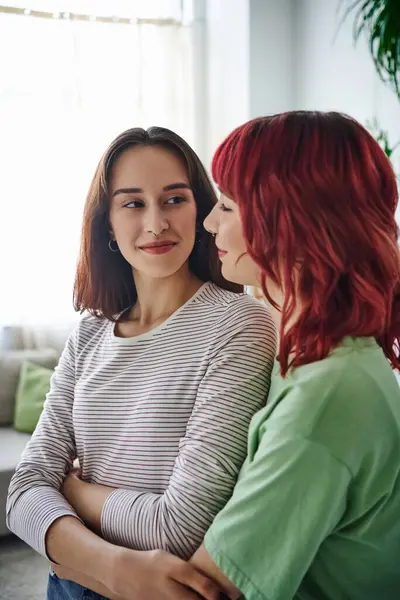 Portrait of cheerful and pierced lesbian woman with red hair looking at her girlfriend at home — Foto stock