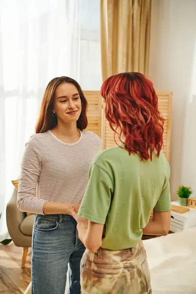 Feliz joven lesbiana de la mano con su novia en apartamento moderno, cariñoso - foto de stock