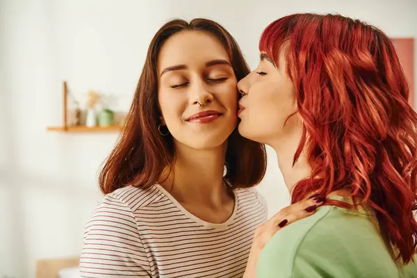 Tendre jeune femme lesbienne aux cheveux roux embrassant la joue de sa petite amie heureuse dans un appartement moderne — Photo de stock