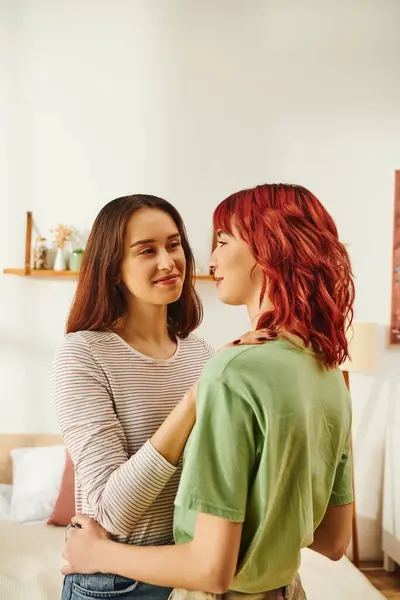 Tierna joven lesbiana abrazando a su novia feliz con el pelo rojo en apartamento moderno - foto de stock