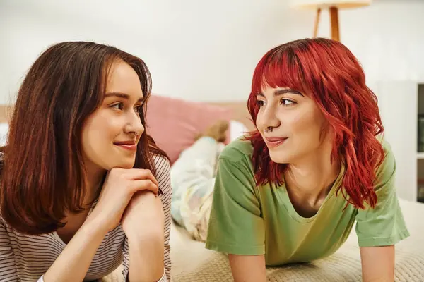 Feliz joven lesbiana pareja en casual atuendo teniendo acogedor momento y mirando el uno al otro en la cama - foto de stock