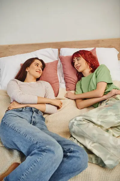 Happy young lesbian couple looking at each other while lying together on bed, domestic life - foto de stock