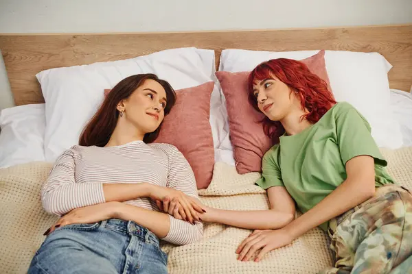 Happy young lesbian couple looking at each other while lying together on bed, holding hands - foto de stock