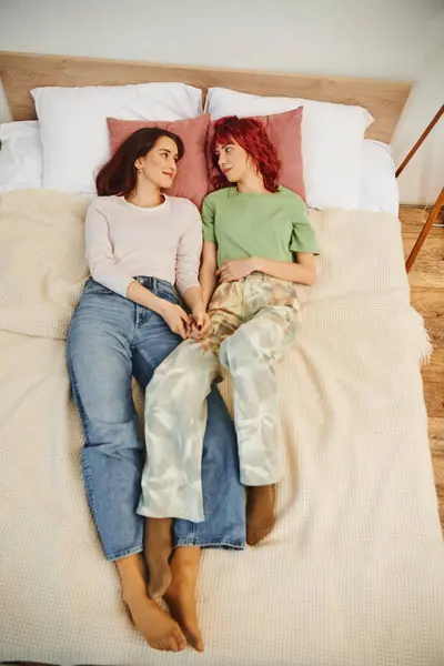 Ansicht von oben: glückliches lesbisches Paar hält sich die Hände, während es zusammen auf dem Bett liegt und einander ansieht — Stockfoto