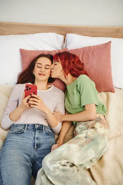 Вид сверху женщины с рыжими волосами, целующей щеку своей подруги со смартфоном, лежа на кровати — стоковое фото