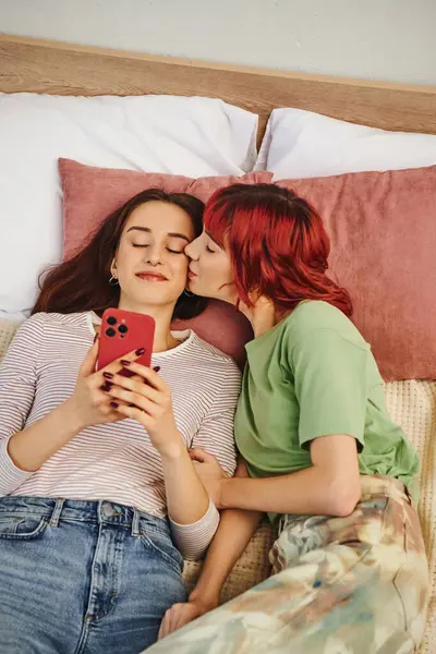 Top view of woman with red hair kissing cheek of her girlfriend with smartphone in hands, bedroom — Stock Photo