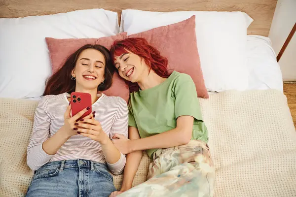 Вид счастливой молодой лесбийской пары, делающей селфи на смартфоне, лежа вместе на кровати — стоковое фото