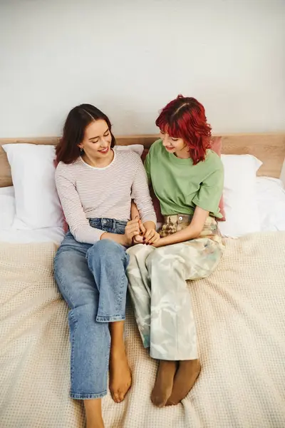 Pleased young lesbian couple in casual wear holding hands while sitting together on bed - foto de stock