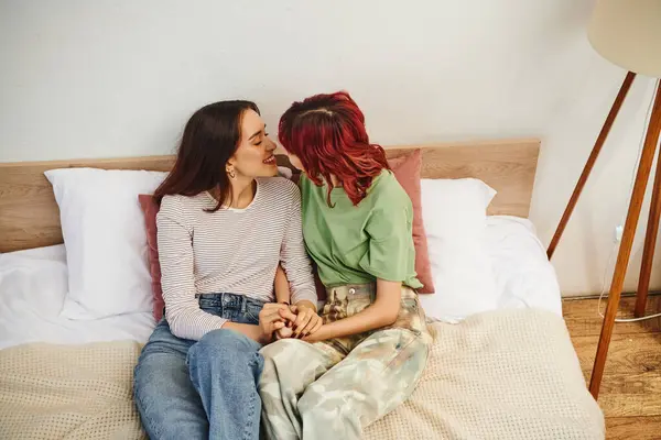 Pleased young lesbian couple in casual wear holding hands and going to kiss while sitting on bed — Stock Photo
