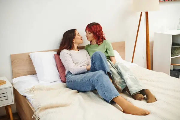 Freudiges junges lgbt-Paar in Freizeitkleidung, das sich beim gemeinsamen Sitzen auf dem Bett ansieht — Stockfoto