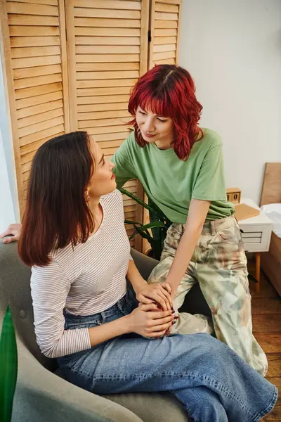Jeune et heureuse femme lesbienne aux cheveux roux regardant sa petite amie assise dans un fauteuil à la maison — Photo de stock