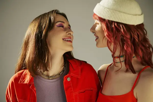 Happy young lgbt couple with bold makeup in vibrant attire looking at each other on grey, lesbian — Stock Photo