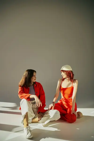 Счастливая стильная лесбийская пара в ярком наряде, сидящая и смотрящая друг на друга в студии на сером — стоковое фото