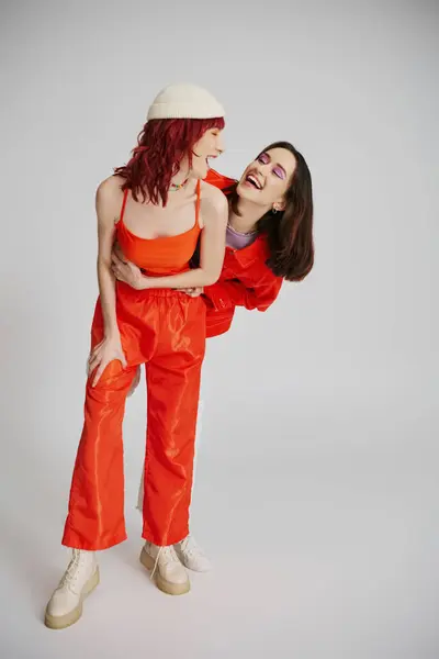 Счастливая стильная лесбийская пара со смелым макияжем в ярком наряде, смотрящая друг на друга на сером — стоковое фото