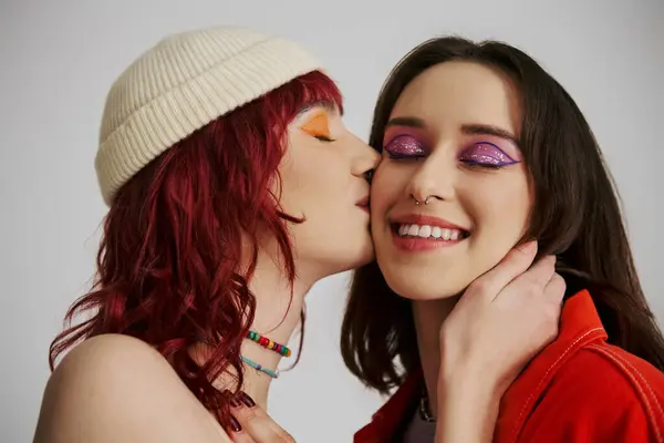 Heureux élégant lesbienne femme avec audacieux maquillage et vibrant tenue baisers joue de copine sur gris — Photo de stock