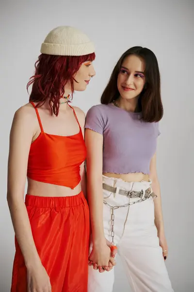 Happy lesbian couple with bold makeup in vibrant attire looking at each other and walking on grey — Stock Photo