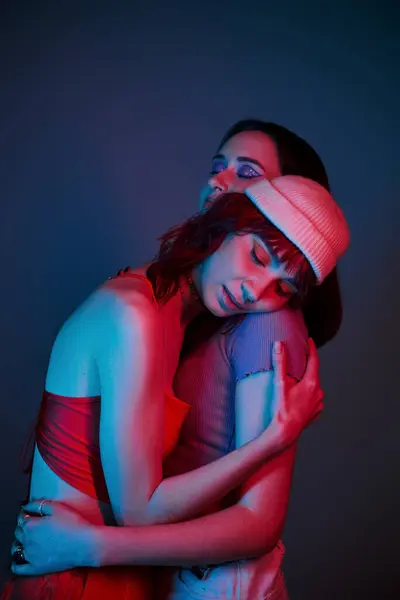 Jeune couple lesbien avec un maquillage audacieux et une tenue élégante étreignant sous des lumières violettes en studio — Photo de stock