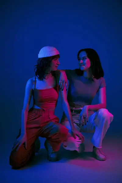 Happy girlfriends in 20s posing in stylish attire and looking at each other under purple lights - foto de stock